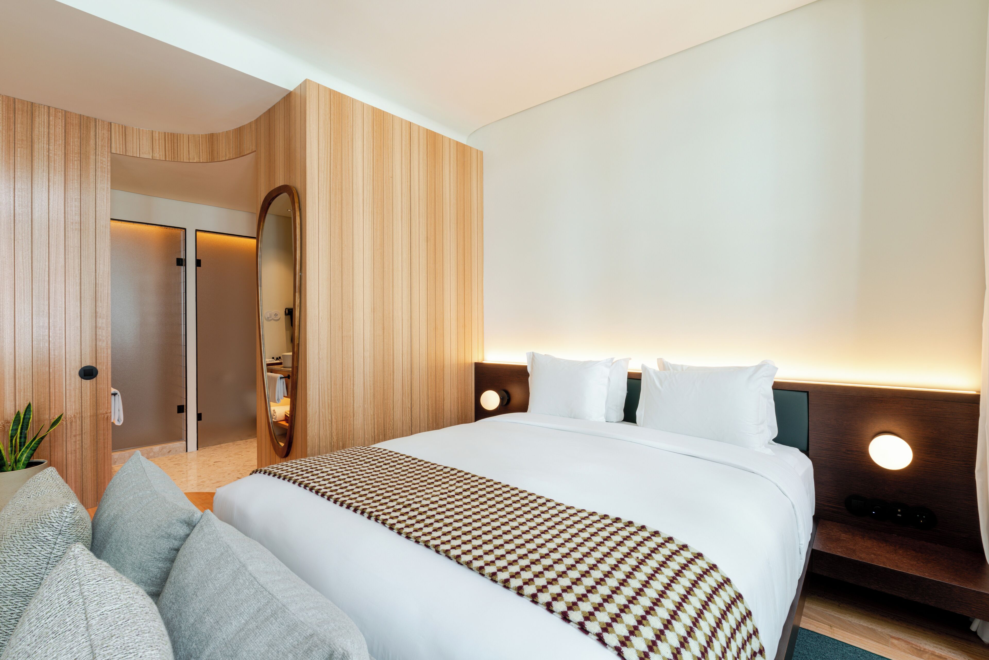 A modern hotel room with a large bed, a wooden accent wall, a mirror, and a bedside table with a lamp.