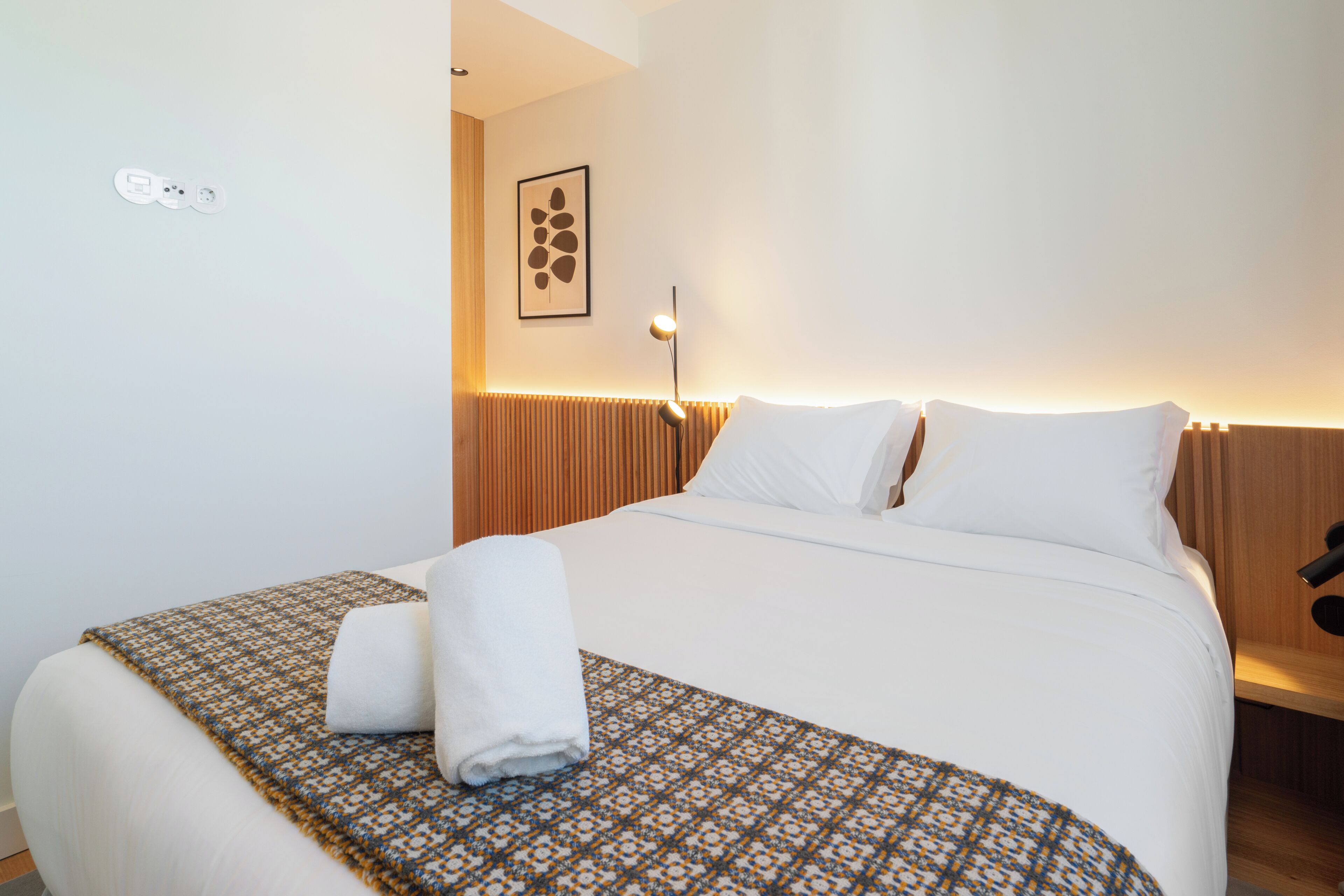 A neatly made bed with a patterned throw, a white towel, and a wall-mounted light fixture.