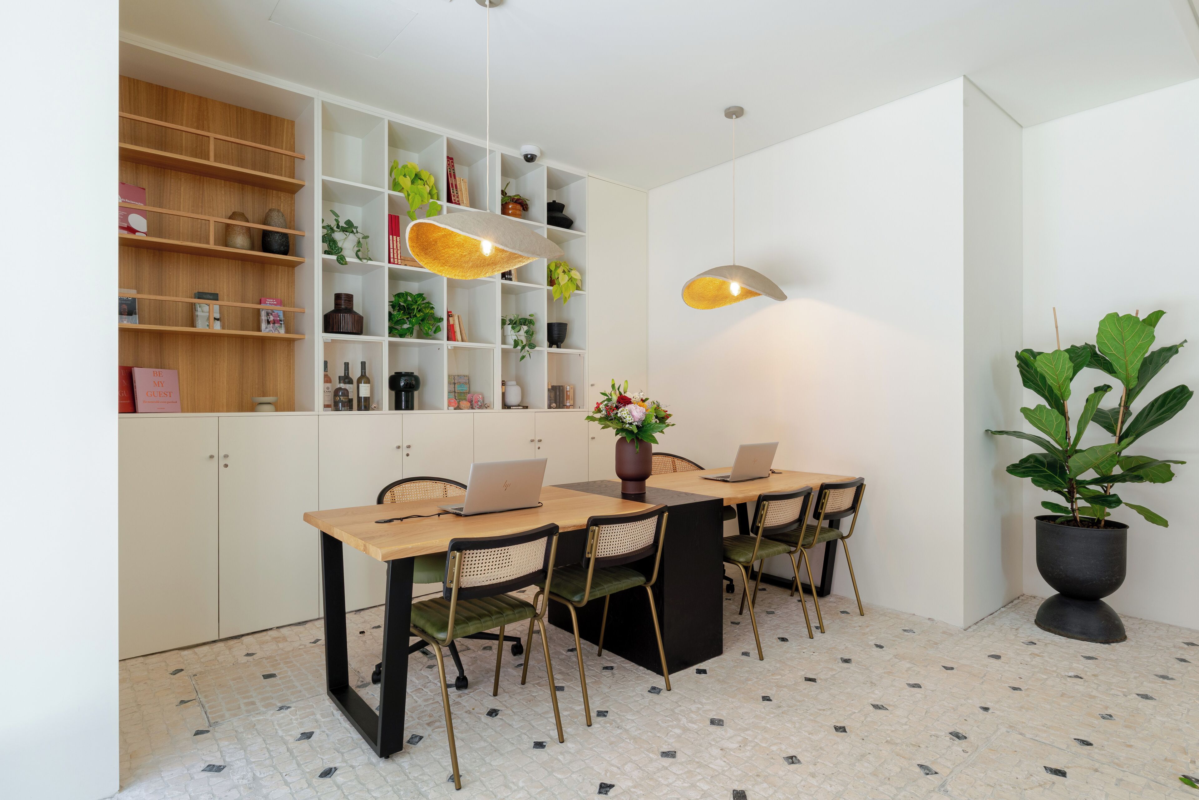 A modern dining area with a wooden table, four chairs, two laptops, a vase with flowers, and a potted plant.