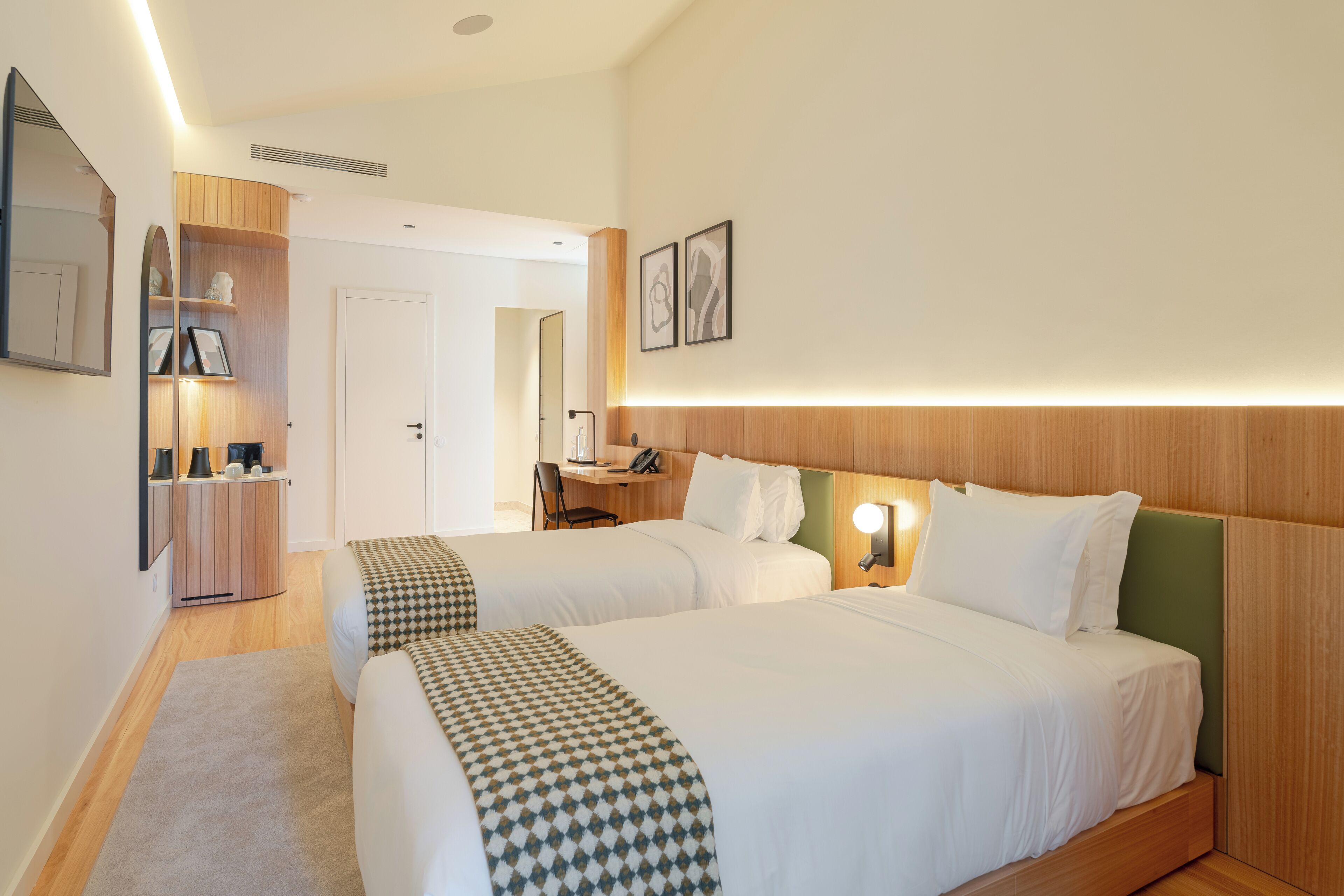 A hotel room with two beds, a desk, a chair, a mirror, and a small shelf with items.