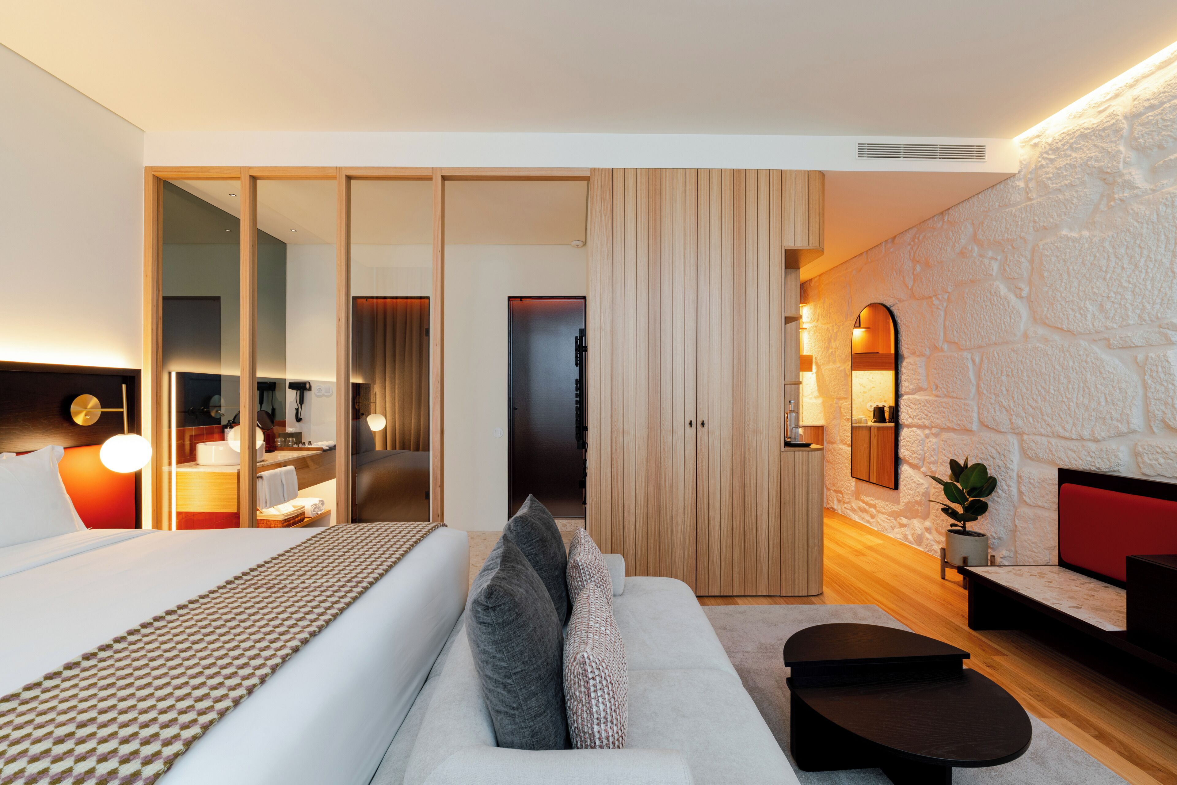 A modern hotel room with a large bed, a sofa, a small table, a wardrobe, and a stone accent wall.
