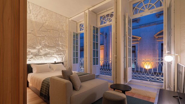 A bedroom with a bed, a sofa, and a small table. There are open doors leading to a balcony with a railing. - NOLIA Boutique Hotel (Porto)