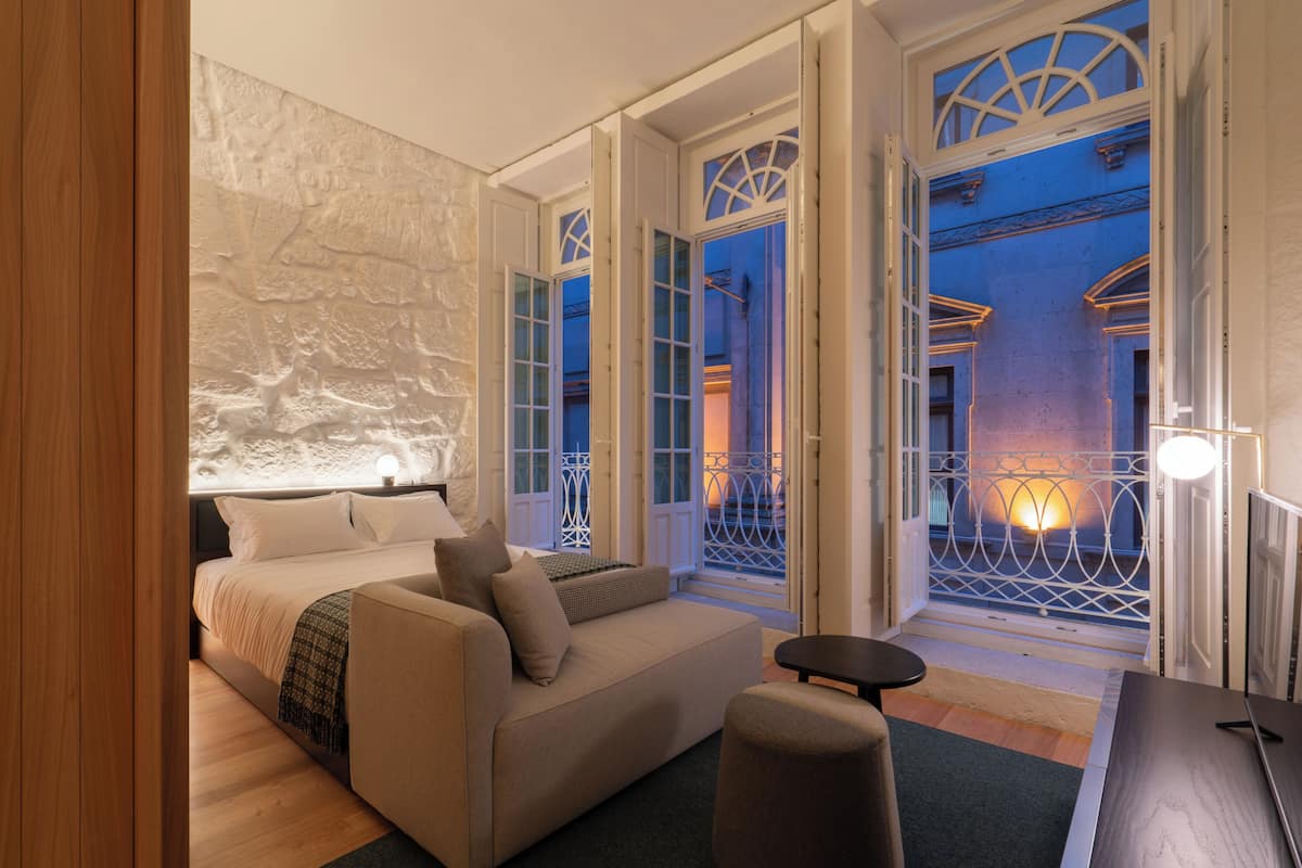 A bedroom with a bed, a sofa, and a small table. There are open doors leading to a balcony with a railing.