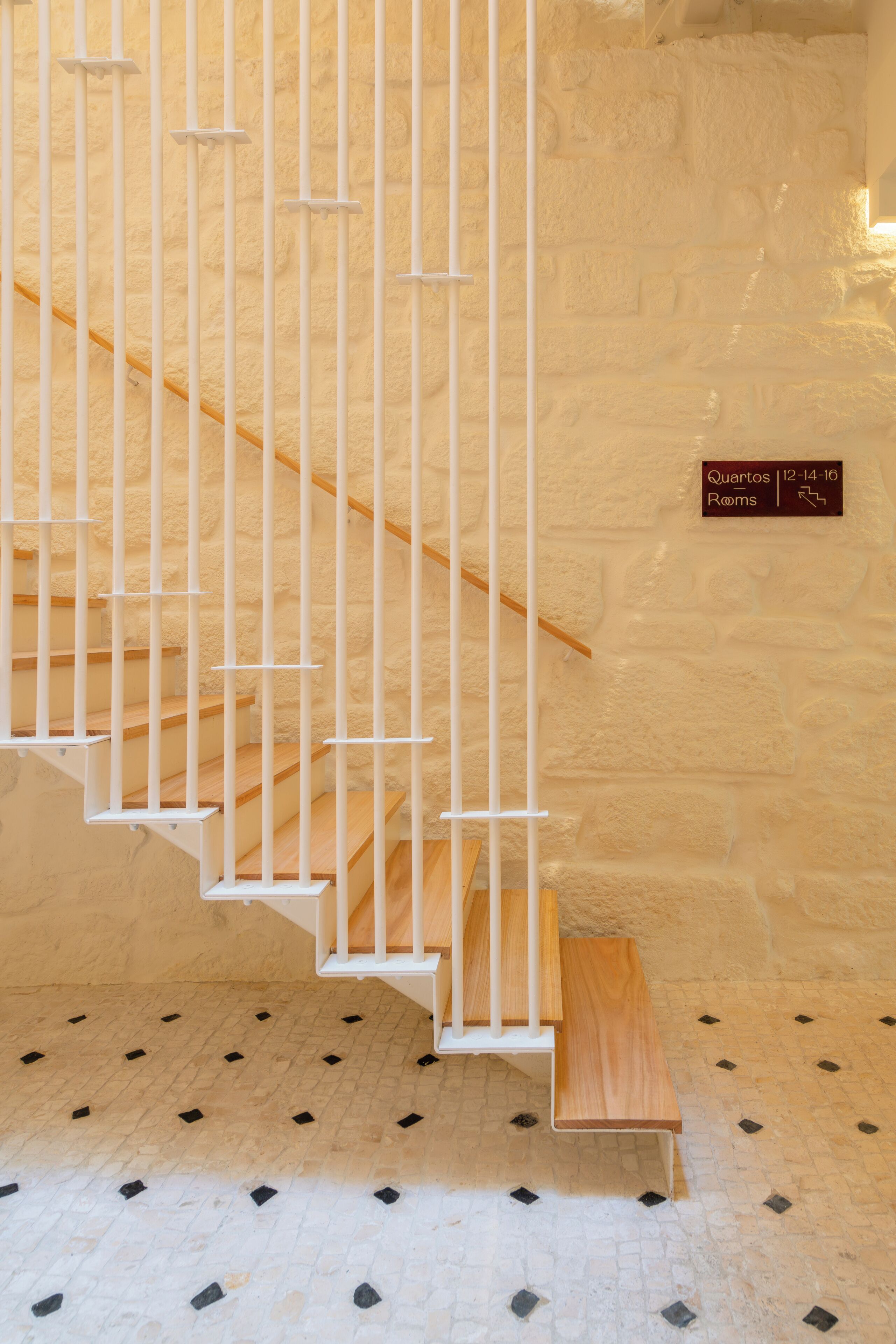 A staircase with white railings and a wooden handrail, leading to a landing with a sign indicating 'Quartos 12-14-16 Rooms'.