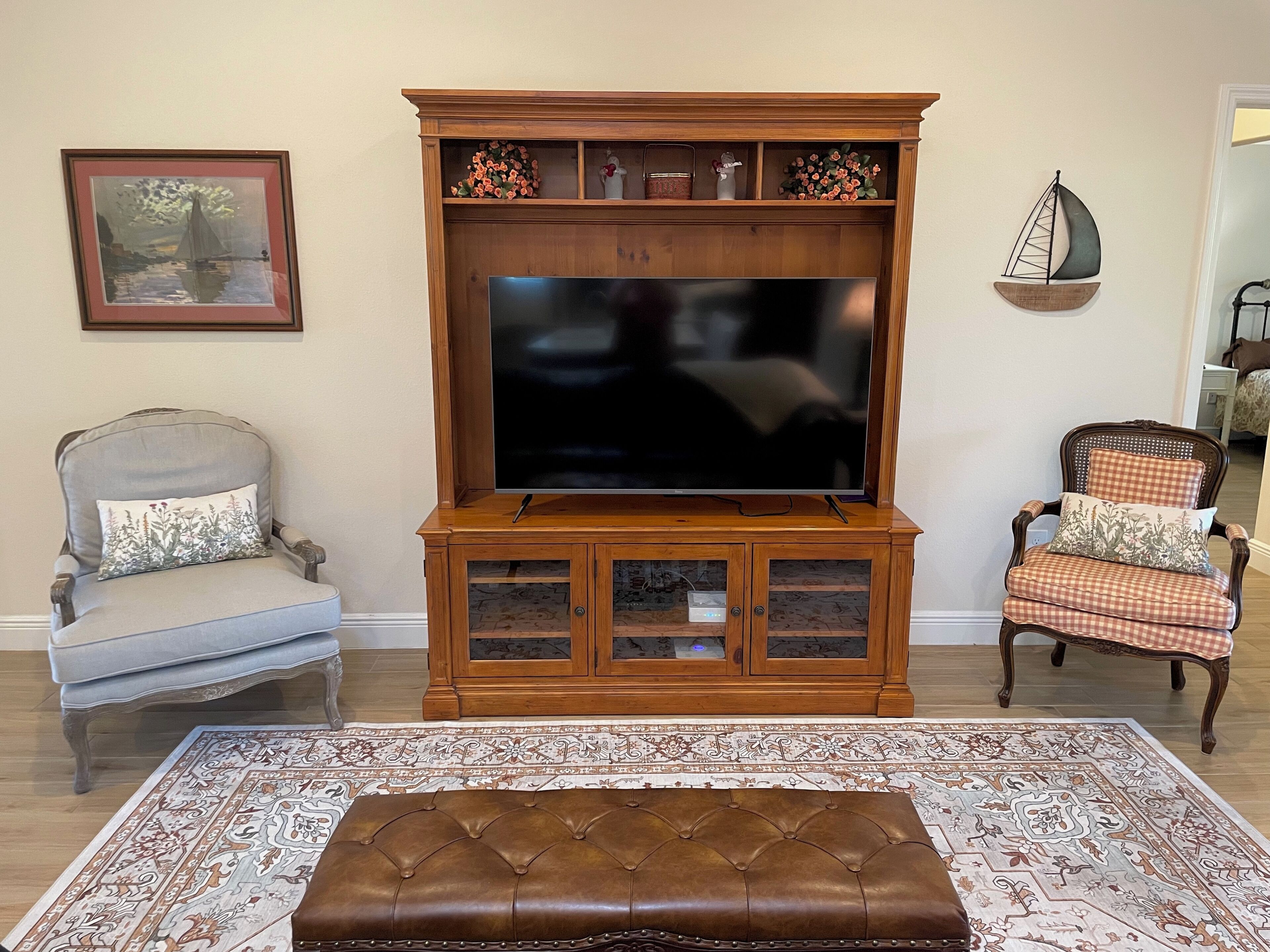 A living room with a large flat-screen TV on a wooden entertainment center, two upholstered chairs, a patterned rug, and a framed picture on the wall.