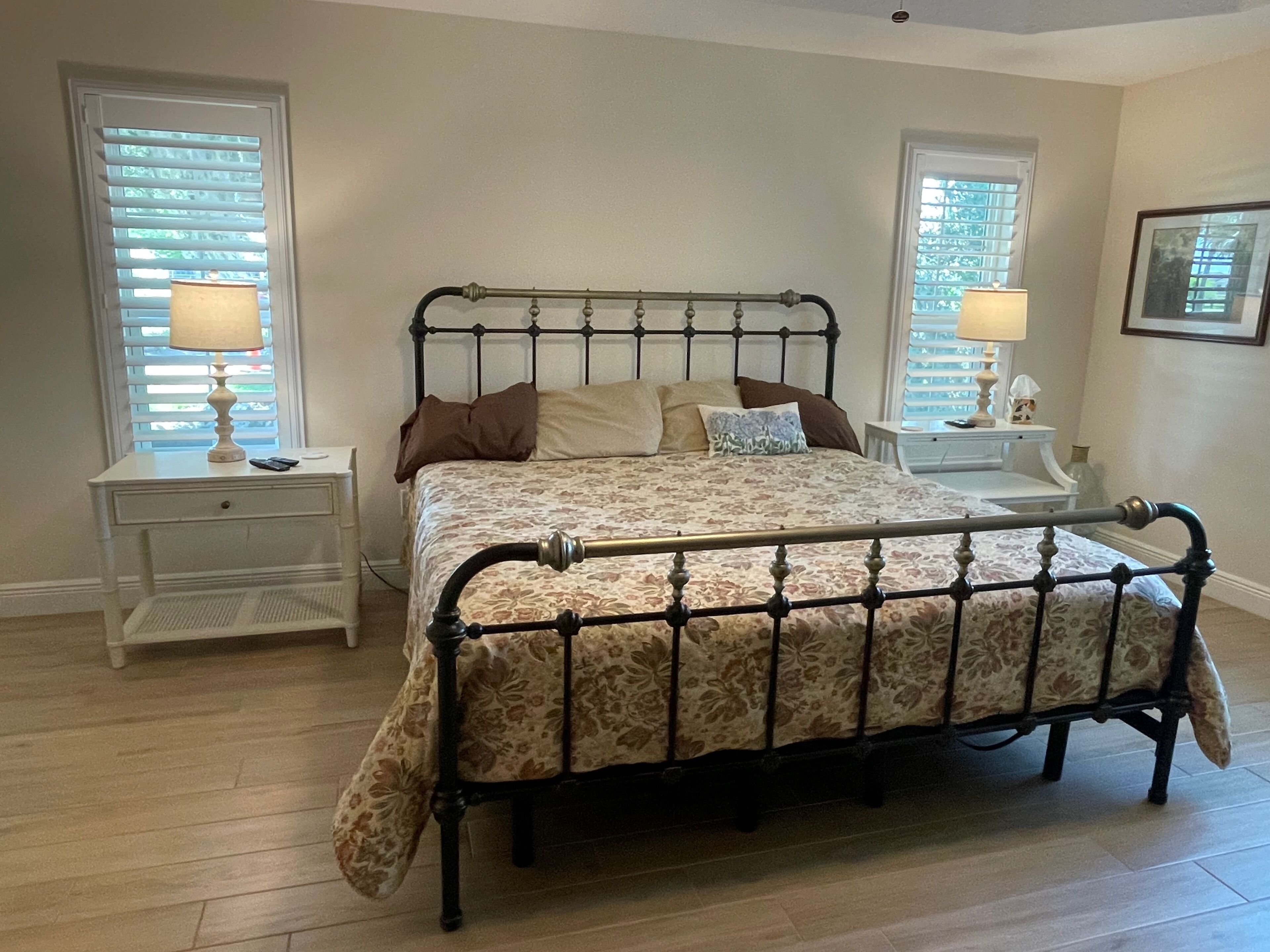 A bedroom with a metal bed frame, two bedside tables with lamps, a white nightstand, and a framed picture on the wall.