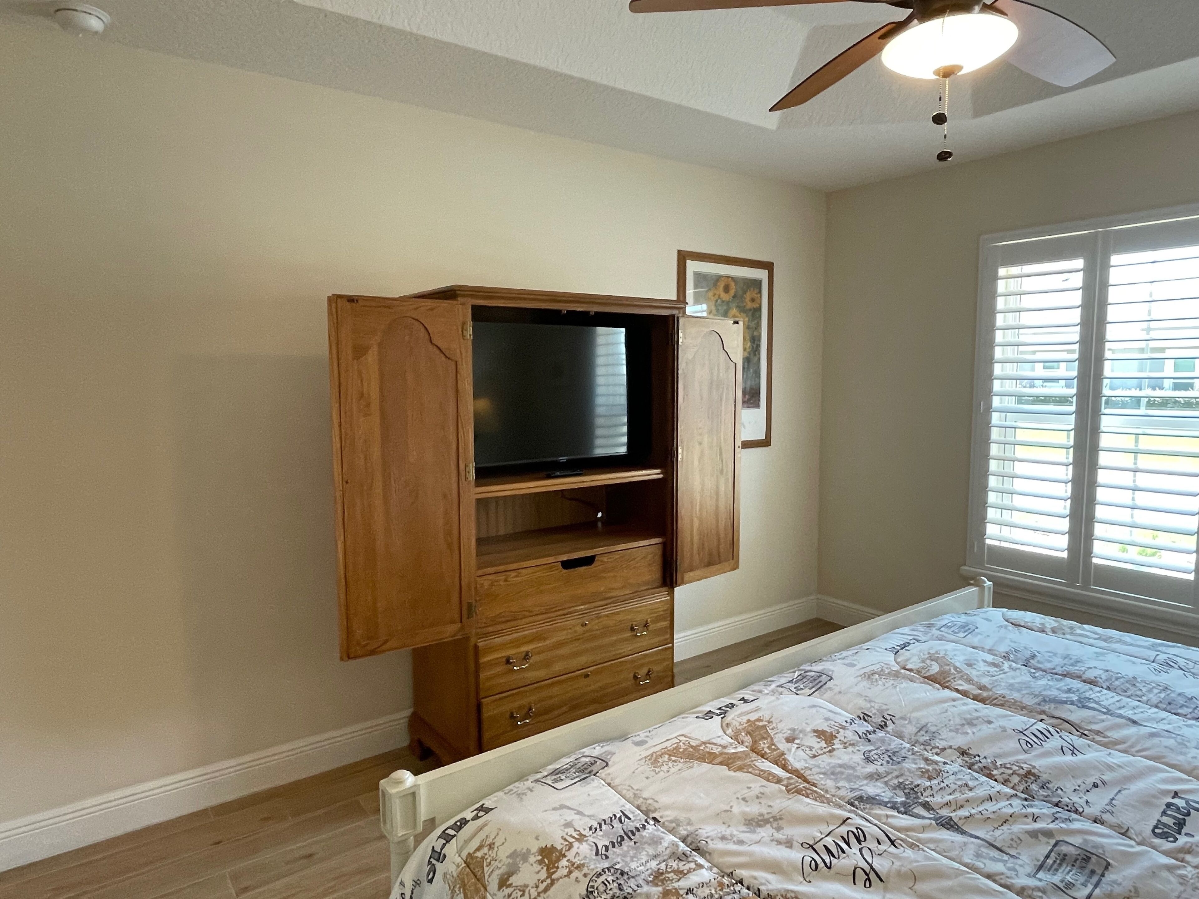 A bedroom with a wooden cabinet, a bed with patterned bedding, a ceiling fan, and a window with blinds.