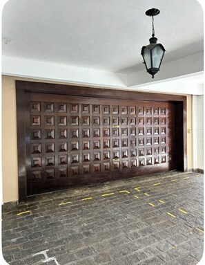 Uma porta de madeira fechada com padrão geométrico, um candeeiro pendurado e um chão pavimentado com marcações amarelas.