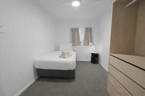 A small, single-bed room with a grey carpet, a bedside table with a lamp, and a window with curtains.