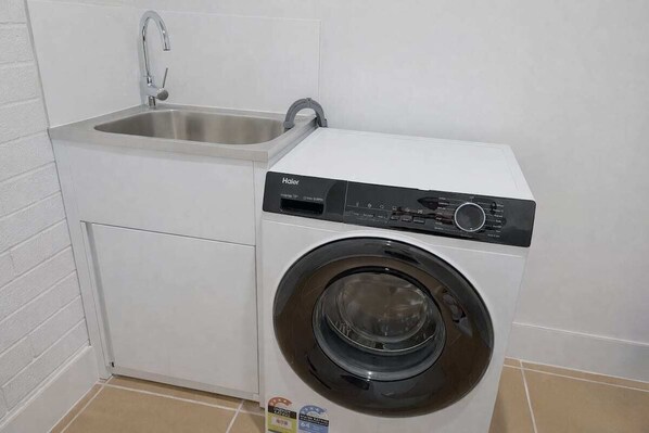 A compact washer and dryer unit with a stainless steel sink, located in a corner of a room with white walls and tiled flooring.