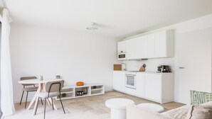 Eine moderne Küche mit weißen Schränken, ein Essbereich mit Tisch und Stühlen sowie ein Wohnbereich mit Sofa und Hocker.
