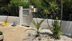 Ein durch eine Mauer abgegrenzter Außenbereich mit weißem Zaun, Palmen und einem Gebäude mit rotem Ziegeldach im Hintergrund.