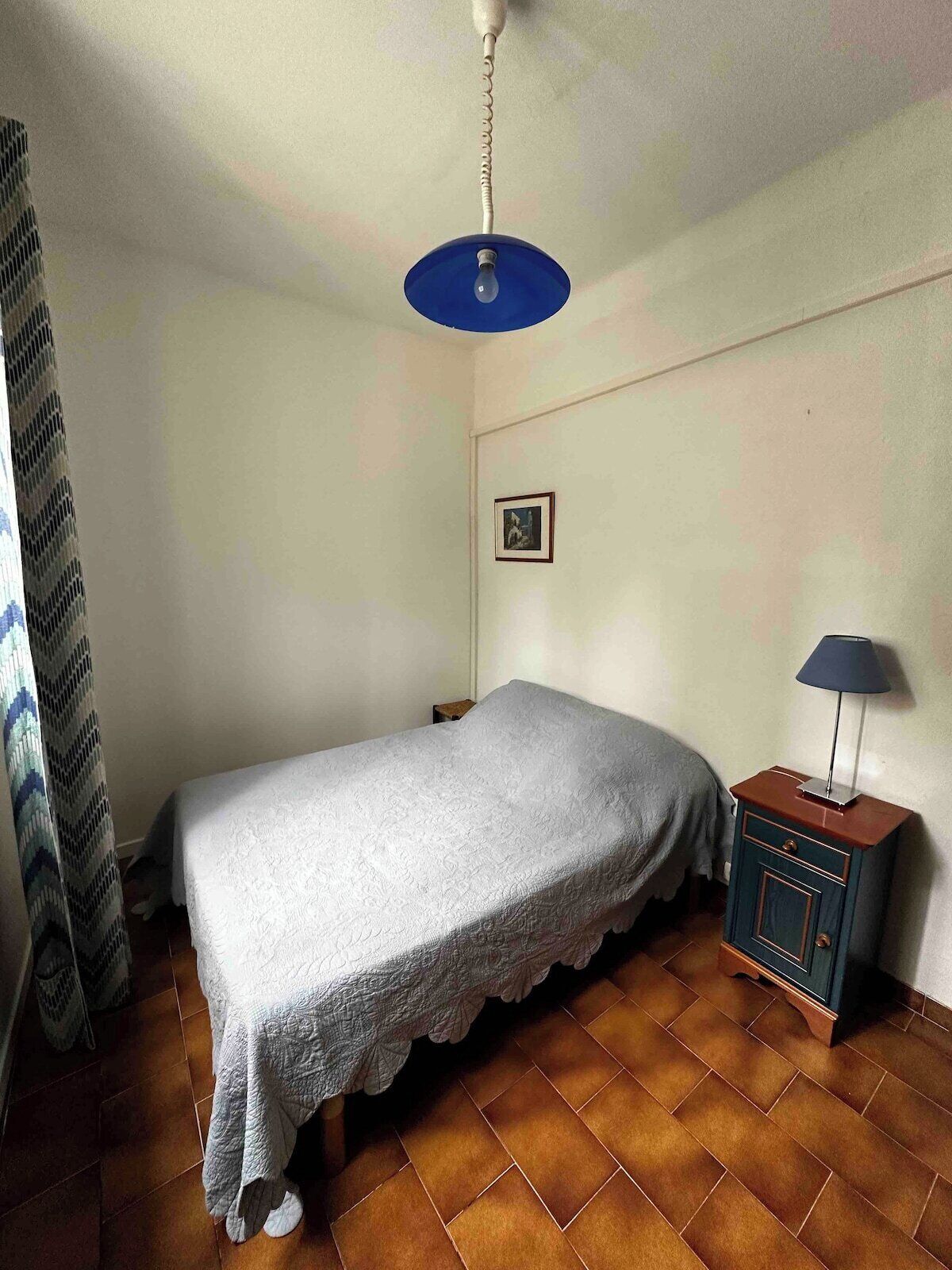 Ein Schlafzimmer mit Bett, Nachttisch mit Lampe, einem Bild an der Wand und einer Deckenleuchte.