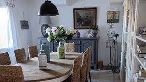 Uma sala de jantar com uma mesa redonda, cadeiras de vime, um vaso com flores, um abajur e um quadro na parede.