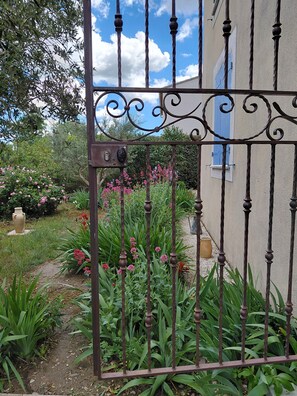 Una reja de hierro forjado con un diseño decorativo, un jardín con diversas plantas y flores, y un edificio con contraventanas azules al fondo.