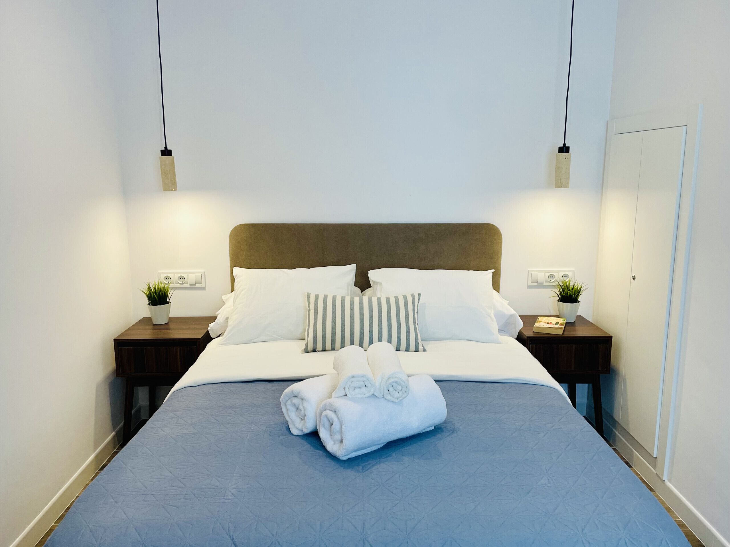 A neatly made bed with a blue bedspread, white pillows, and rolled towels. There are two bedside tables with small potted plants and a nightstand with a lamp.
