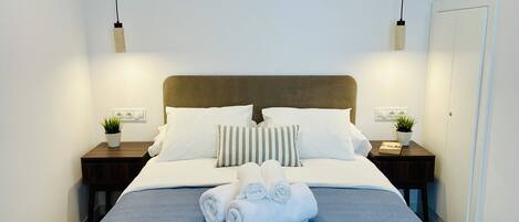 A neatly made bed with a blue bedspread, white pillows, and rolled towels. There are two bedside tables with small potted plants and a nightstand with a lamp.