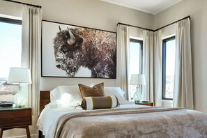 Ein Hotelzimmer mit einem großen Bison-Gemälde, einem Bett mit weißen Leinen, zwei Nachttischlampen und Blick durch die Fenster.