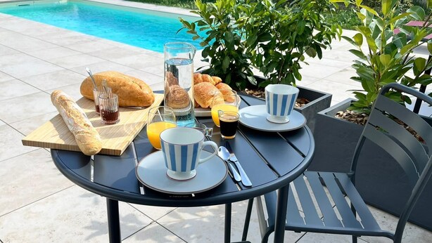 Uma mesa redonda ao ar livre com um jarro de vidro, croissants, uma baguete e uma xícara de café.