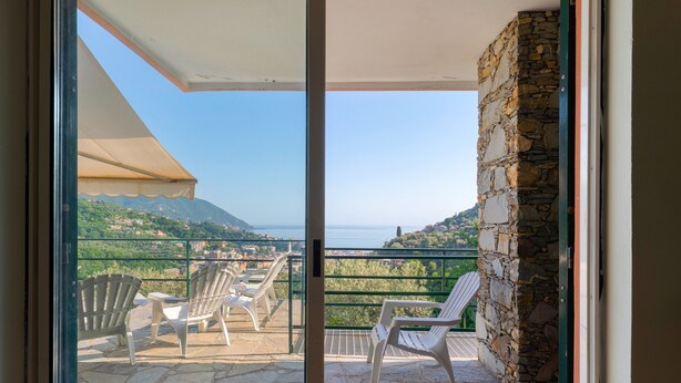 A balcony with chairs, a stone wall, and a view of the sea and mountains.