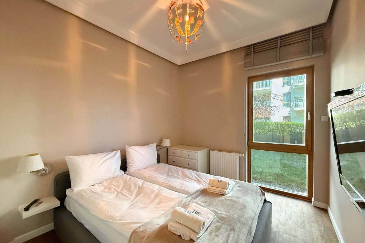 Une chambre d’hôtel moderne avec un grand lit, des tables de chevet, une lampe, un radiateur et une fenêtre donnant sur un paysage verdoyant.