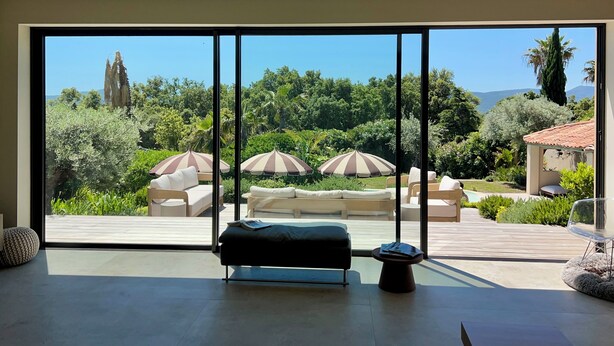 Ein modernes Zimmer mit einer großen SchiebeglasTür, die auf eine Terrasse mit Liegestühlen und Sonnenschirmen führt.