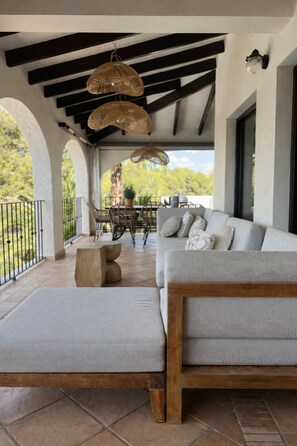 Un espacio al aire libre cubierto con una zona de estar que incluye un sofá, un reposapiés, una mesa, sillas y lámparas de mimbre colgantes.