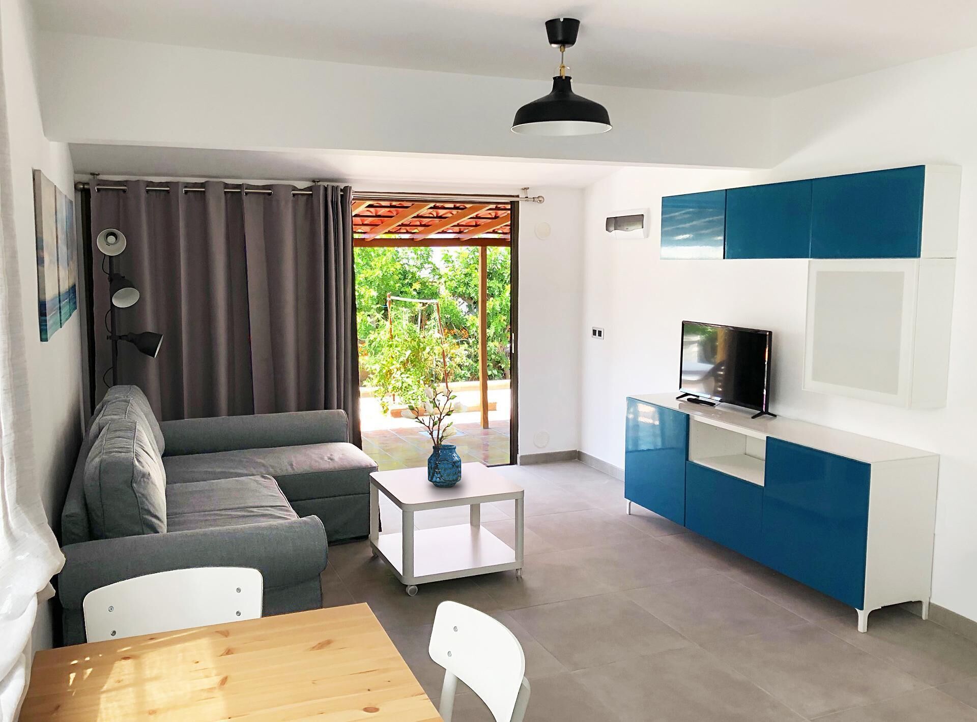 Una sala de estar moderna con un sofá, una mesita, un televisor en un mueble azul y una puerta corrediza de vidrio que da a un jardín.