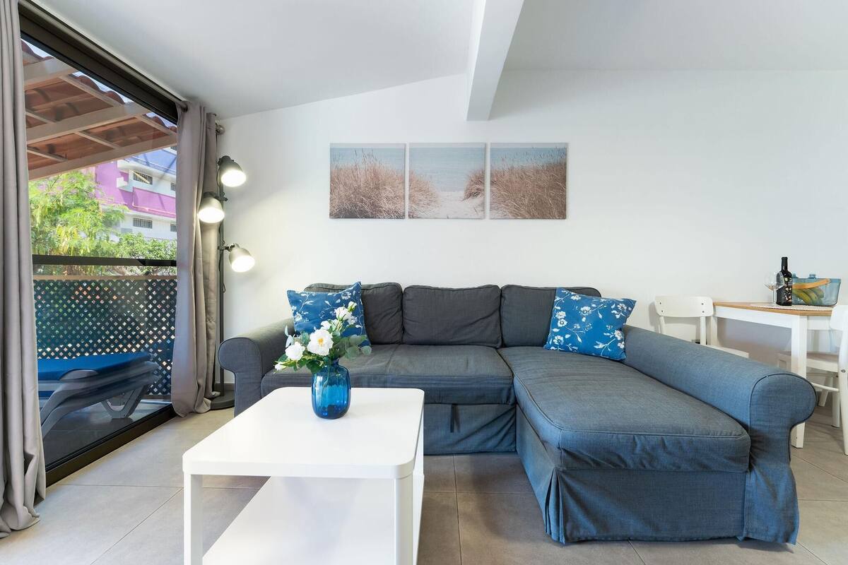 A modern living room with a grey sectional sofa, a white coffee table, and a blue vase with flowers. There is a window with curtains, a wall-mounted picture, and a white table with a bottle.