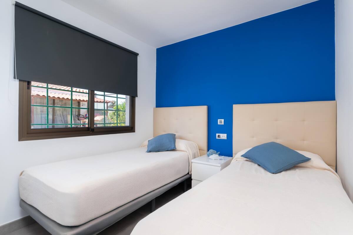 A room with two beds, a window with a blind, and a blue accent wall.