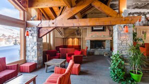 Un intérieur en bois avec des murs en pierre, des meubles rembourrés de couleur rouge, une cheminée et une vue sur des maisons enneigées.