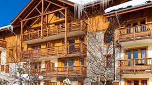Un chalet en bois à plusieurs étages, doté de balcons, entouré d’un paysage enneigé et surplombé d’un ciel bleu clair.