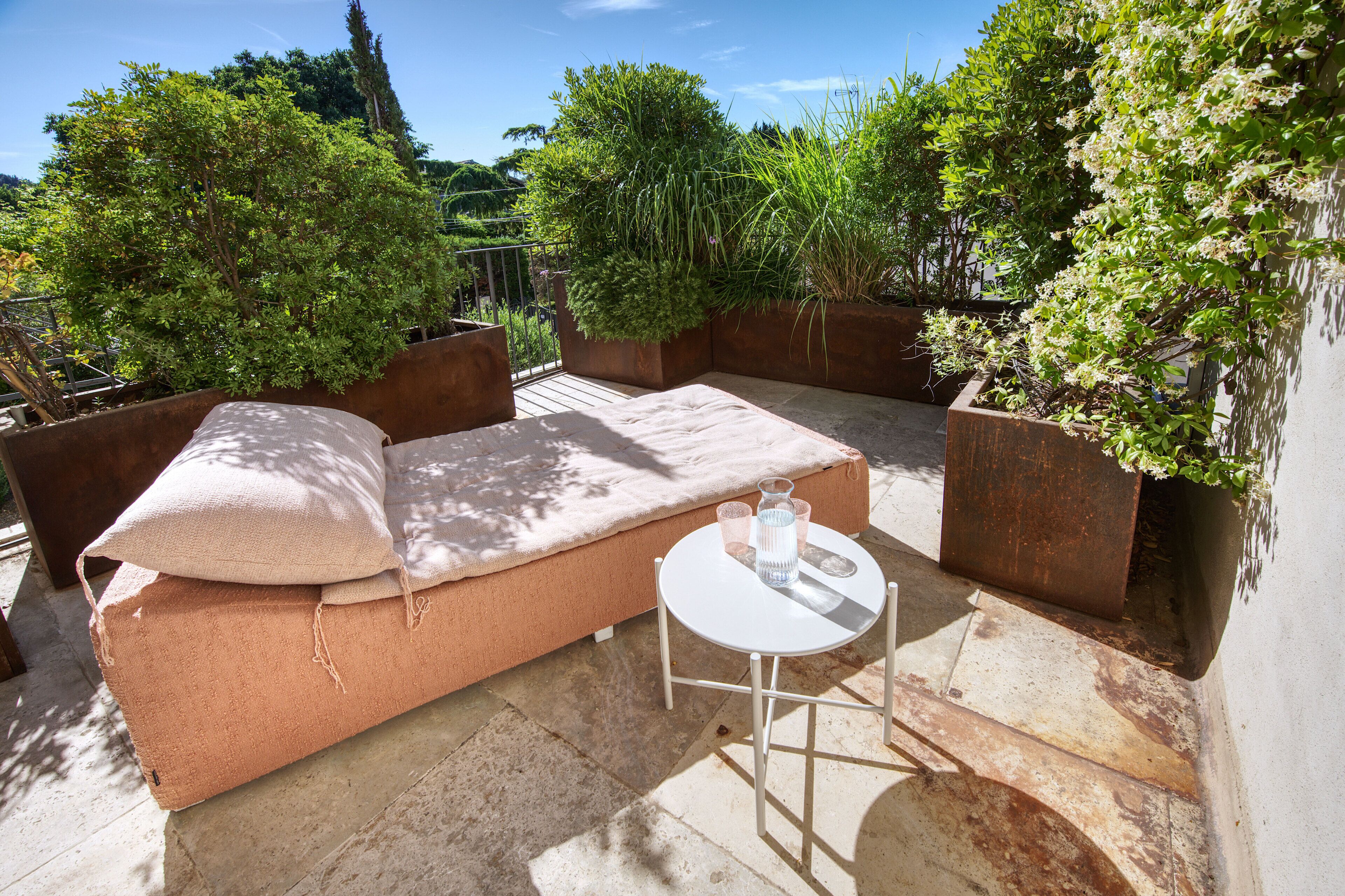 A rooftop patio with a cushioned seating area, a small table with a glass, and potted plants.