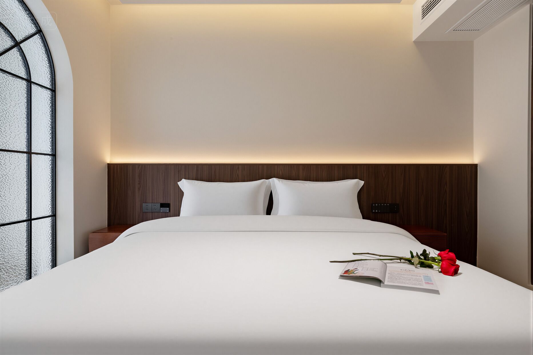 A modern hotel room with a large bed, a wooden headboard, and a bedside table with a book and a rose.