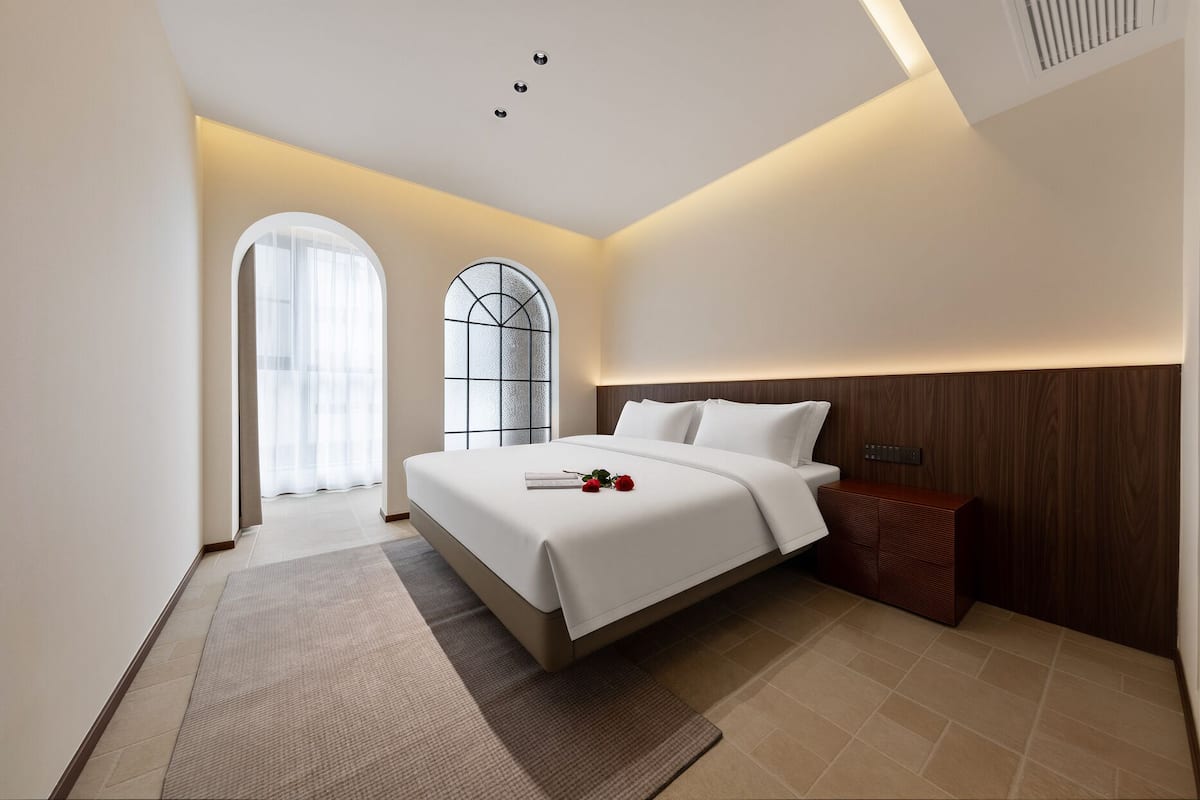Una habitación de hotel moderna con una cama grande, una mesita de noche, un cabecero de madera, un ventanal con vistas y una alfombra pequeña.