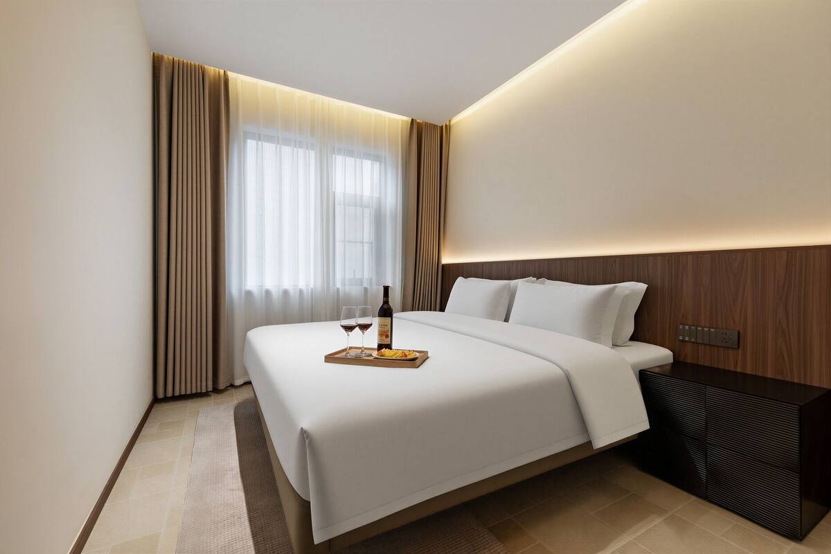 Habitación de hotel con una cama grande, una mesita de noche con una botella y vasos, y una bandeja con comida.