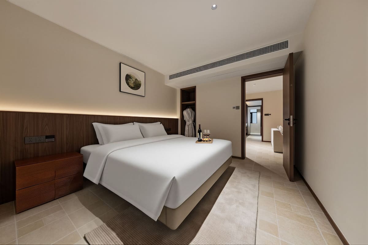 Una habitación de hotel moderna con una cama grande, cabecero de madera, una mesita de noche, una bata colgada en un perchero y una bandeja con una botella y vasos.