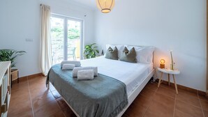 Quarto com cama, mesa de cabeceira com abajur, janela com cortinas e vista para o verde lá fora.