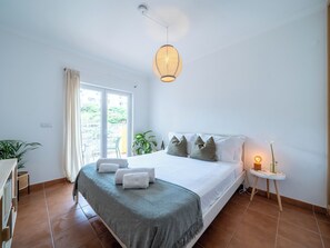 Ein Schlafzimmer mit Bett, Nachttisch mit Lampe, Fenster mit Vorhängen und Blick ins Grüne.