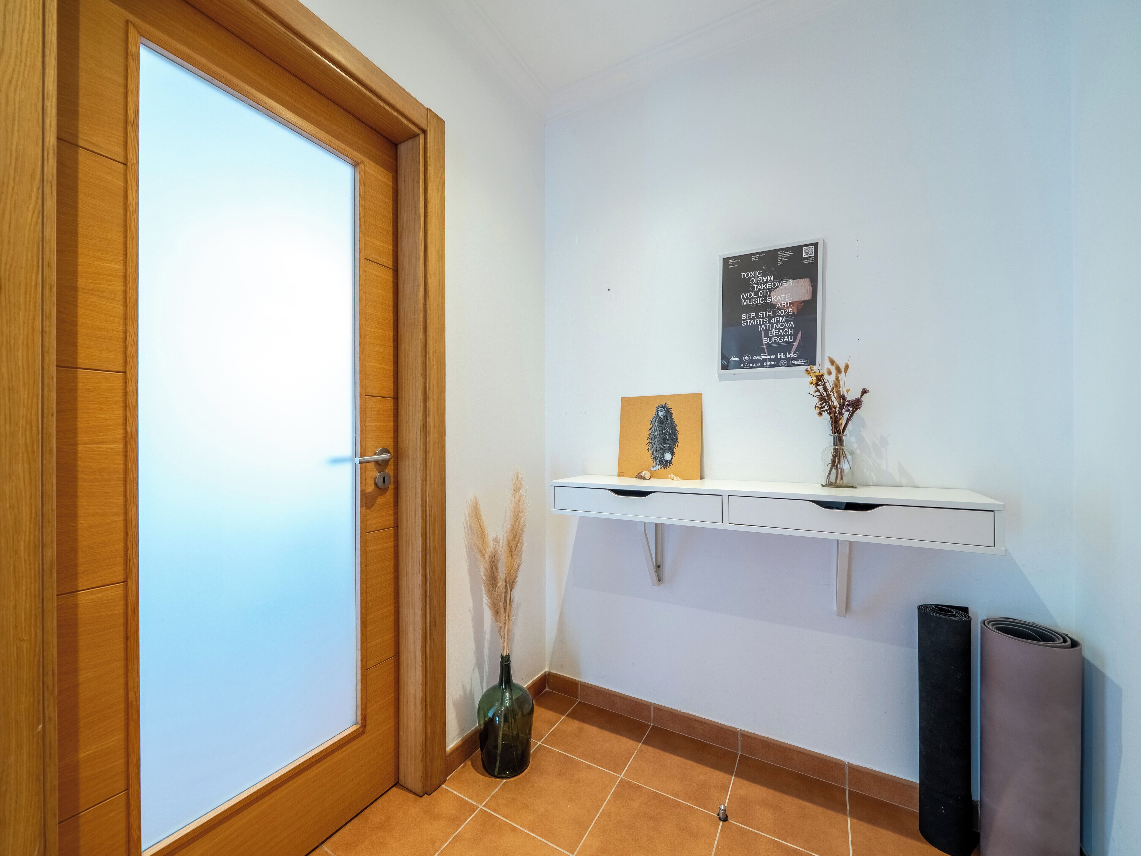 Una habitación con puerta de madera, pared blanca, repisa con un cuadro enmarco, un jarrón con plantas secas y dos esteras enrolladas.