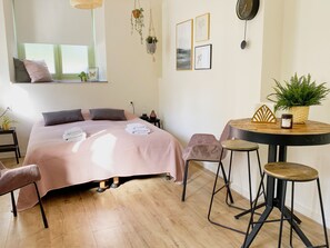 Um quarto moderno com cama, mesas de cabeceira, uma mesa de jantar redonda com cadeiras, uma planta pendente e um relógio de parede.