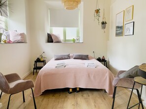 Um quarto com uma cama, duas mesas de cabeceira, uma janela, uma planta pendente e um painel com quadros.