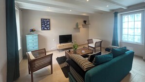 A living room with a sofa, a coffee table, a TV, a bookshelf, and a window. - 4 P. house in a charming Breton village (Pont-Aven)