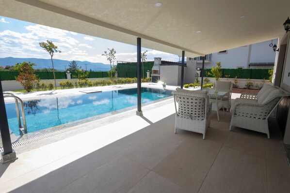 A poolside area with a view of mountains, outdoor furniture, and a covered patio.