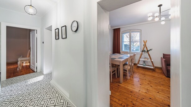 Un pasillo con suelo de diseño geométrico, una zona de comedor con mesa y sillas de madera, y una ventana con cortinas.