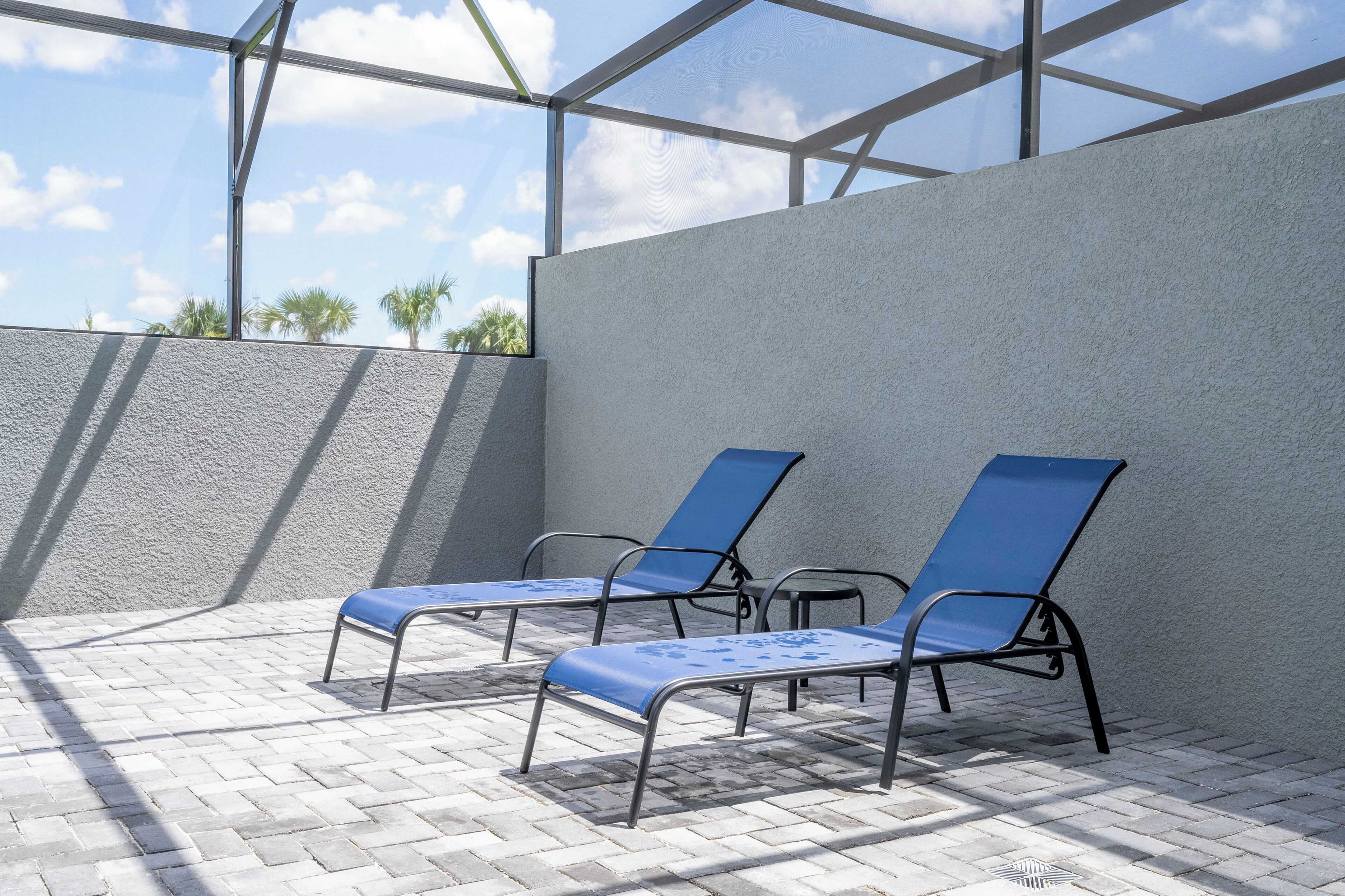 A covered outdoor area with two blue lounge chairs and a small table, featuring a clear view of palm trees and a blue sky through a glass ceiling.