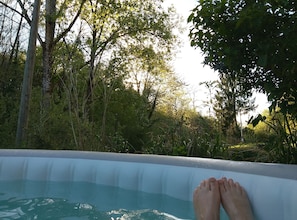 Une piscine dont on aperçoit les pieds d’une personne au bord, entourée d’une végétation luxuriante et d’arbres.