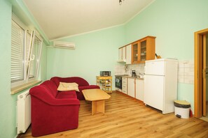 A compact living space with a red sofa, a small wooden table, a kitchen area with cabinets and appliances, and a radiator. - Apartment with balcony and parking space for up to 4 people (Crikvenica)