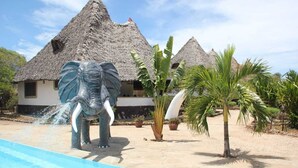 Ein Pool mit Wasserspiel, ein Gebäude mit Reetdach und eine Palme.