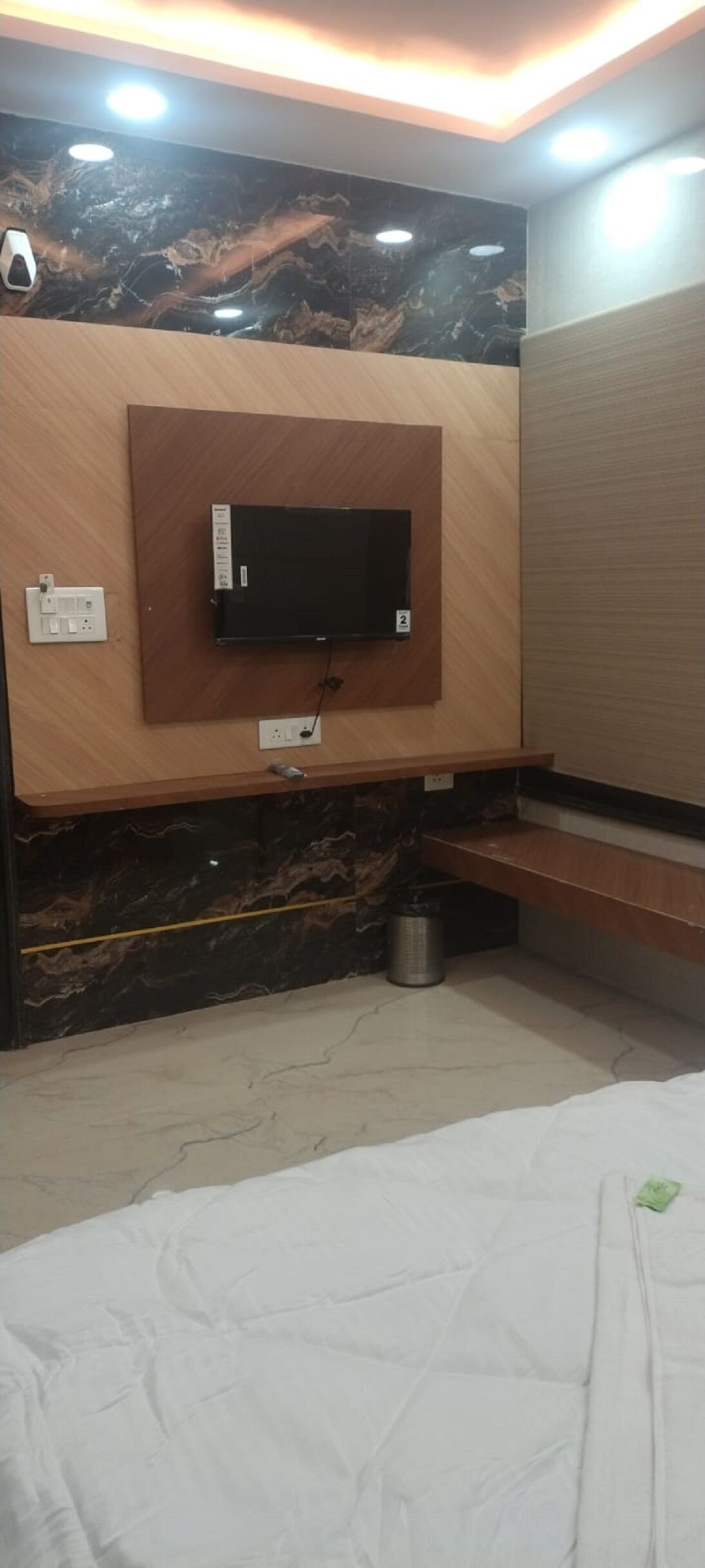 A hotel room with a flat-screen TV mounted on a marble wall, a wooden bench, a trash bin, and a bed with a white mattress.